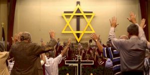 congregation viewed from back of room, many with hands raised; on front wall is a large brown wooden cross with a large yellow Jewish star in front of it