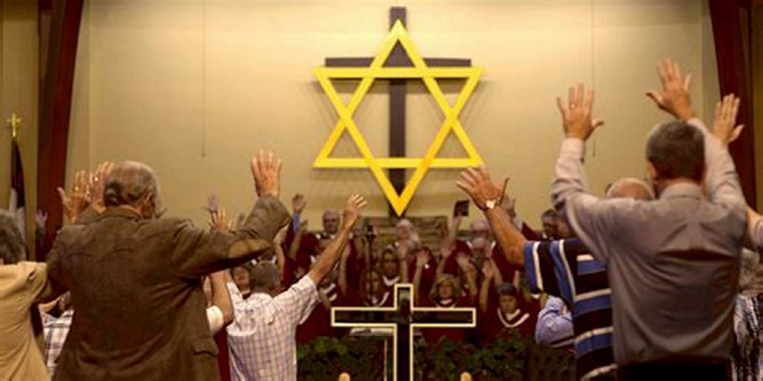 congregation viewed from back of room, many with hands raised; on front wall is a large brown wooden cross with a large yellow Jewish star in front of it