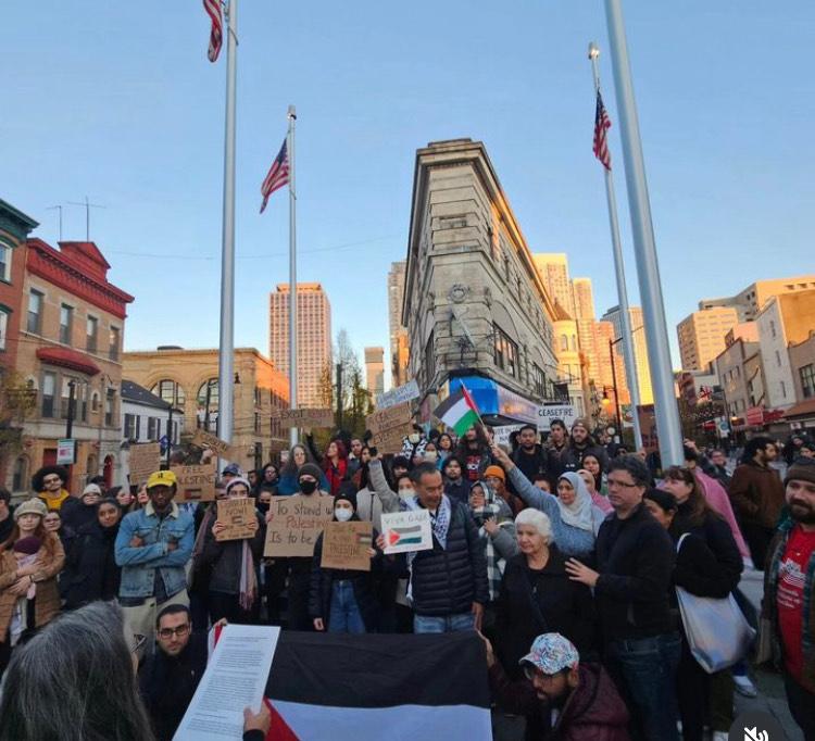 packed crowd at rally on a sunny day in Pedestrian Plaza, Jersey City; people holding cardboard signs, Palestinian flag being waved by woman in hijab; speaker’s notes visible at bottom of photo