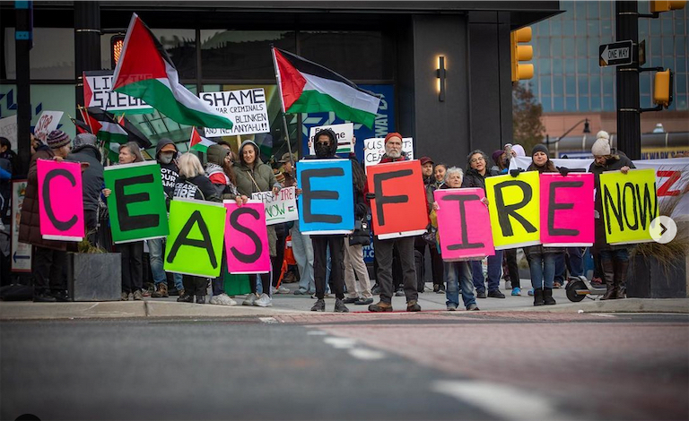 Ralliers in Newark with Palestinian flags, front row holding posters of single letters spelling CEASEFIRE, last one holding the word Now.