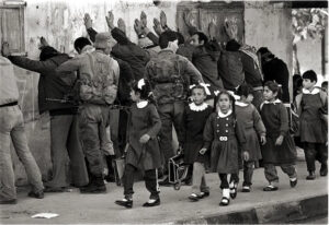 1960s-era black-and-white photo of elementary-school children walking past soldiers holding guns controlling a group of men facing a wall with their hands against the wall above their heads