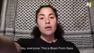 Young woman with dark hair and eyes in front of a keffiah wears a black shirt with collar and lapel microphone. She is reaching out toward screen as though to turn on a TV; caption reads “Hey, everyone. This is Bisan from Gaza.” Upper right corner has logo for AJ+.