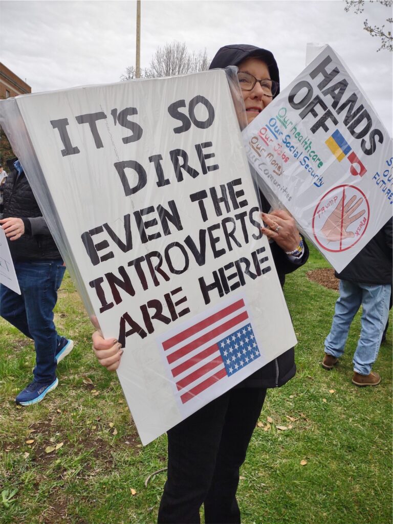 Woman in hooded jacket and classes holds two signs; the more prominent has the message “IT’S SO DIRE EVEN THE INTROVERTS ARE HERE” above an upside-down U.S.A. flag (signalling distress). The second sign has a large HANDS OFF with 2 flags (Ukraine and a red-and-white flag), and a list of items: our healthcare, our Social Security, our jobs, our money, and our communities.