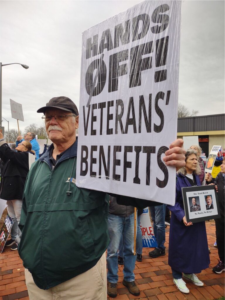 Older gentleman in cap and windbreaker holding a sign with the message “HANDS OFF! VETERANS’ BENEFITS”; more ralliers and signs in the background.
