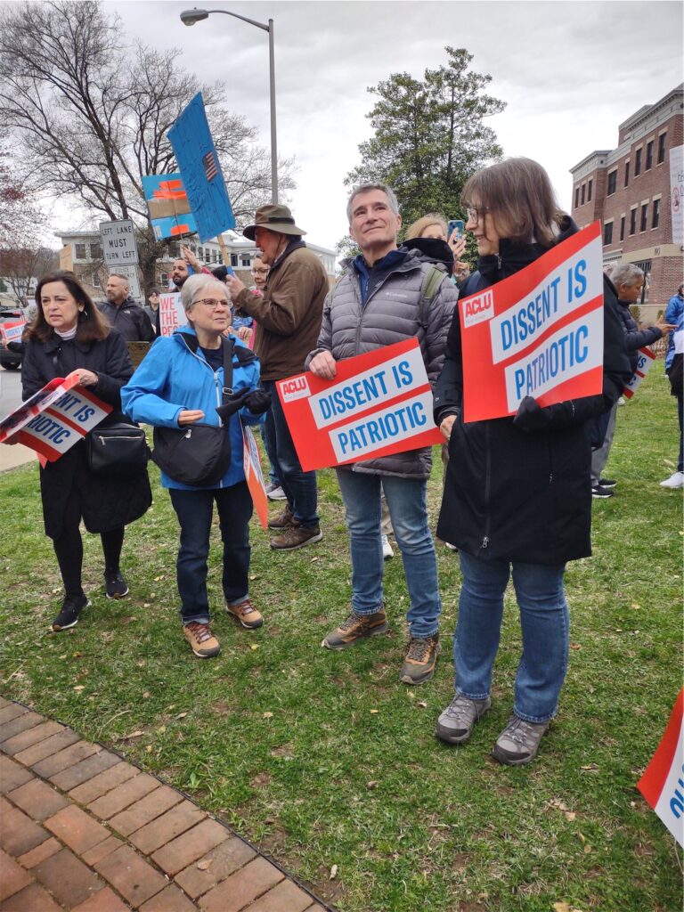 Ralliers on grass holding ACLU signs with the message “DISSENT IS PATRIOTIC”, with additional ralliers and signs in the background.