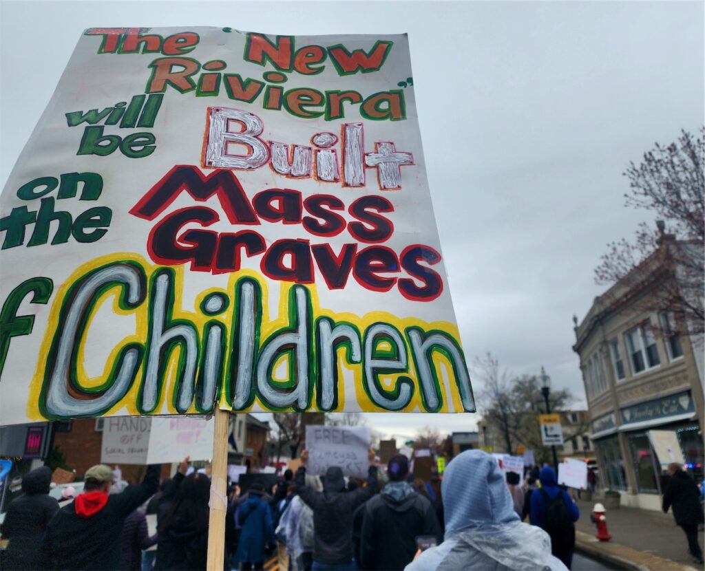 The focus is a large homemade sign with the message “The ‘New Riviera’ will be Build on the Mass Graves of Children”. Visible marching ahead are a crowd of people holding other homemade HandsOff signs.