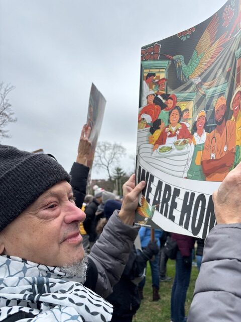 Man in a keffiyeh holds an immigrant-supporting sign that says “WE ARE HONORED”.