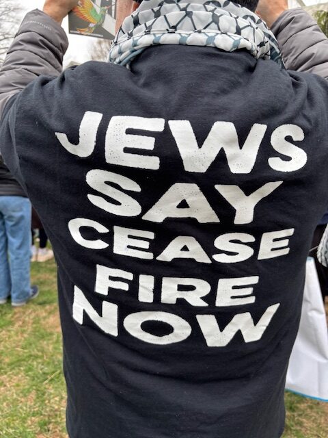 Close-up of the back of a man wearing T-shirt with the message “JEWS SAY CEASE FIRE NOW”, wearing a keffiyeh around his neck