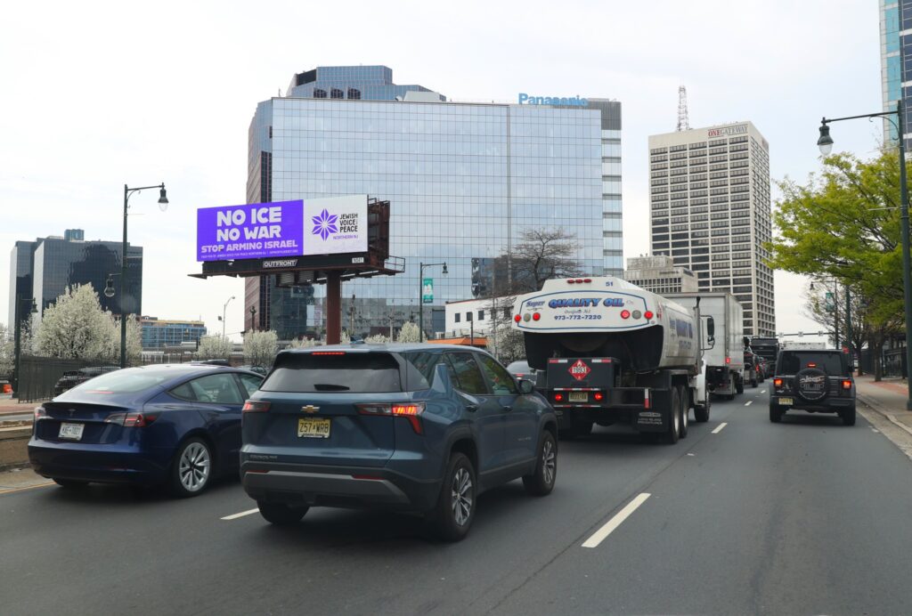 Billboard in Newark is seen in context, wtih cars driving past, mirrored high-rise building behind, and surrounding urban buildings and trees