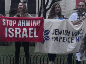 Activists hold cloth banners reading, in white capital letters on red, “Stop Arming Israel” and, in purple letters on white, with a JVP star logo, “Jewish Voice for Peace NYC”.