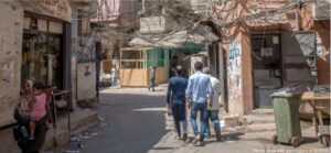 Palestinian refugees walk in littered street of refugee camp with graffiti, wires and ropes overhead, overfull trash can, appearing shabby and temporary